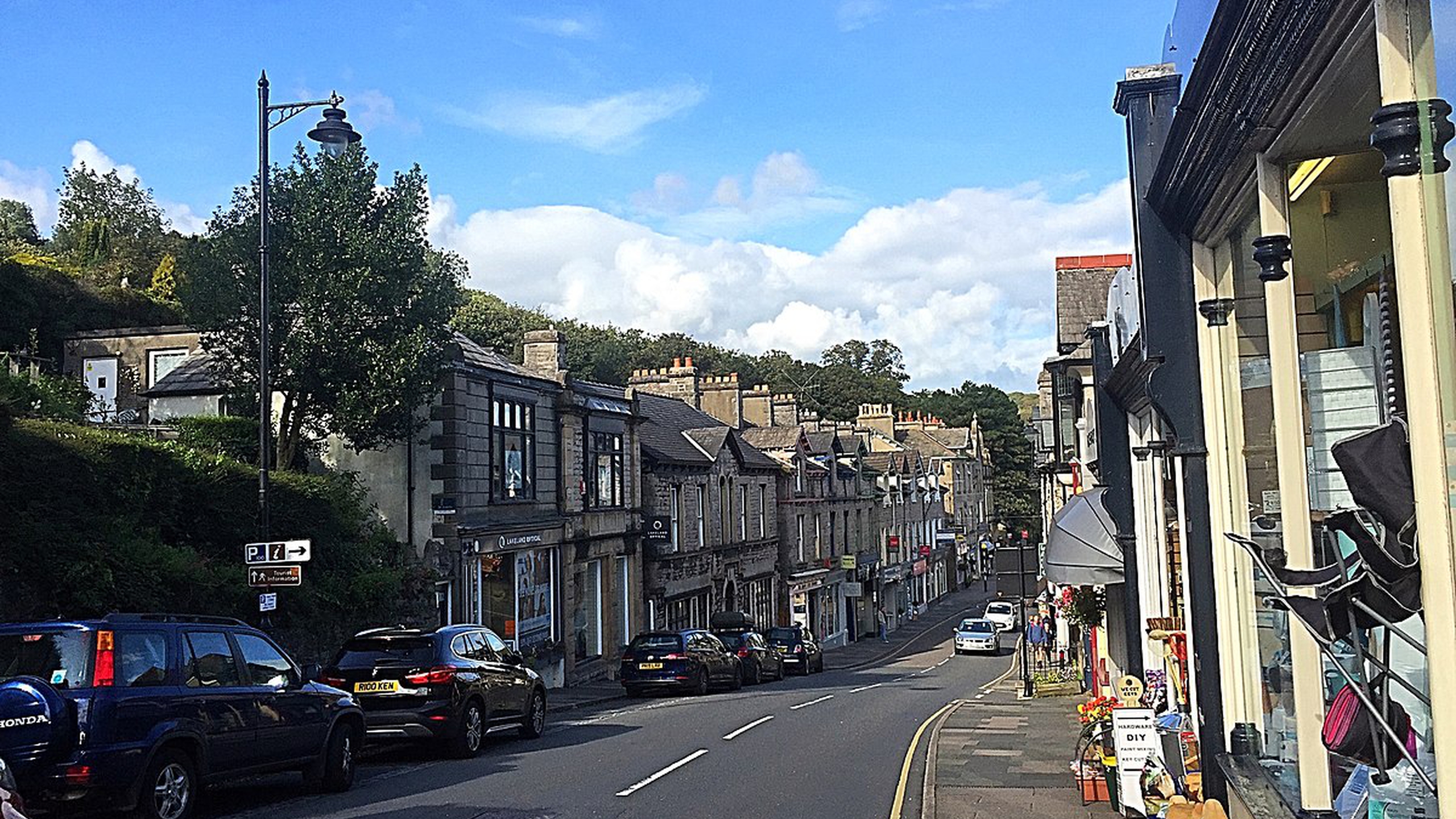A photograph of Main Street, Grange Over Sands, taken from the top of the street on a bright sunny day, showing expensive cars and handsome old buildings.