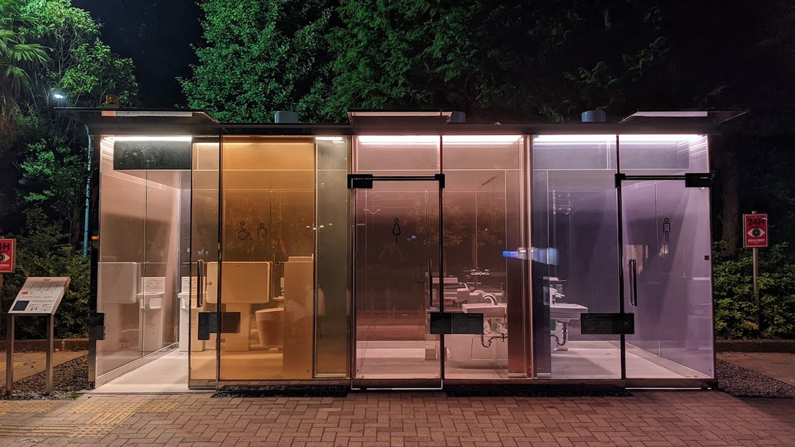 A photograph of a public toilet in Tōkyō, taken at night. The toilet building is made of coloured glass in shades of blue and orange, so is transparent, and is glowing slightly in the streetlamps.